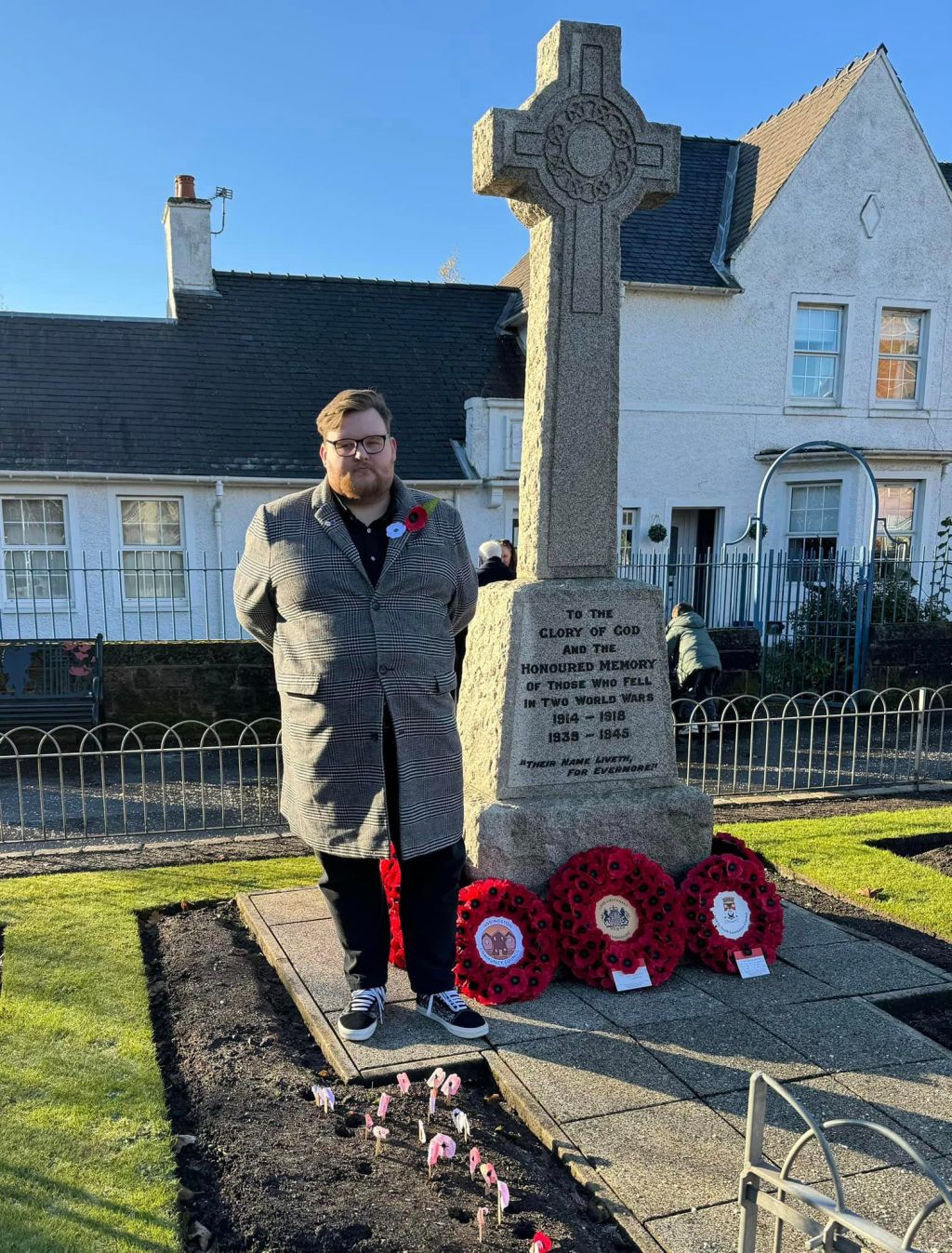 Cllr Cal Johnston-Dempsey standing in front of Uddingston War Memorial, wearing a white and a red poppy, wreaths of poppies from the council, community council, and Lanarkshire Lieutenancy are laid,